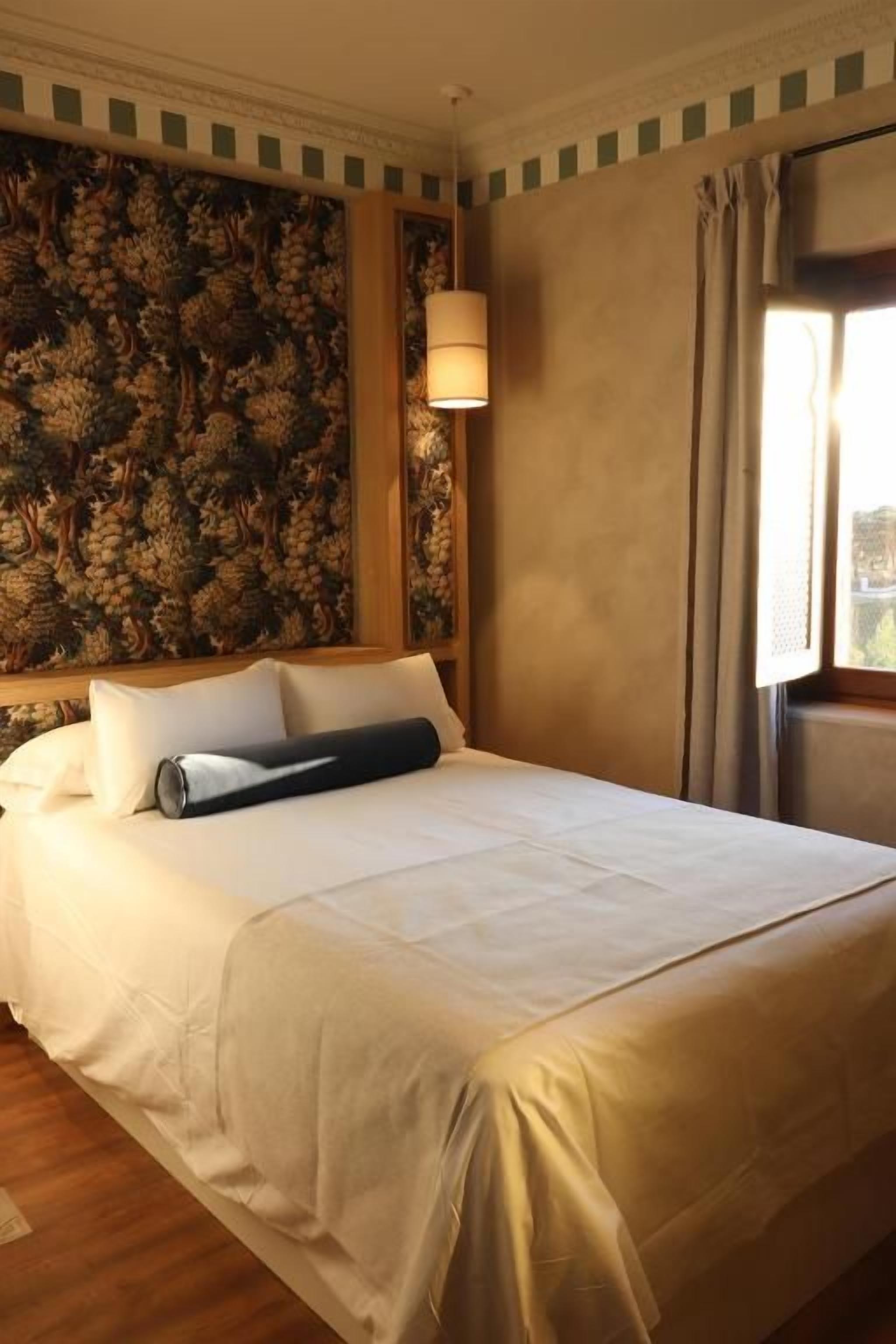 Una habitación de hotel con una cama grande, papel tapiz floral, una ventana con cortinas y una lámpara fijada a la pared.