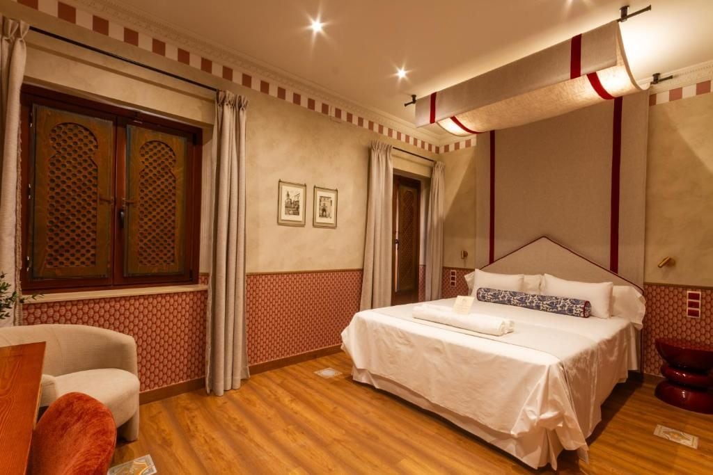 Habitació d'hotel amb un llit, una cadira, un sòl de fusta, una finestra amb cortines i una paret amb quadres.