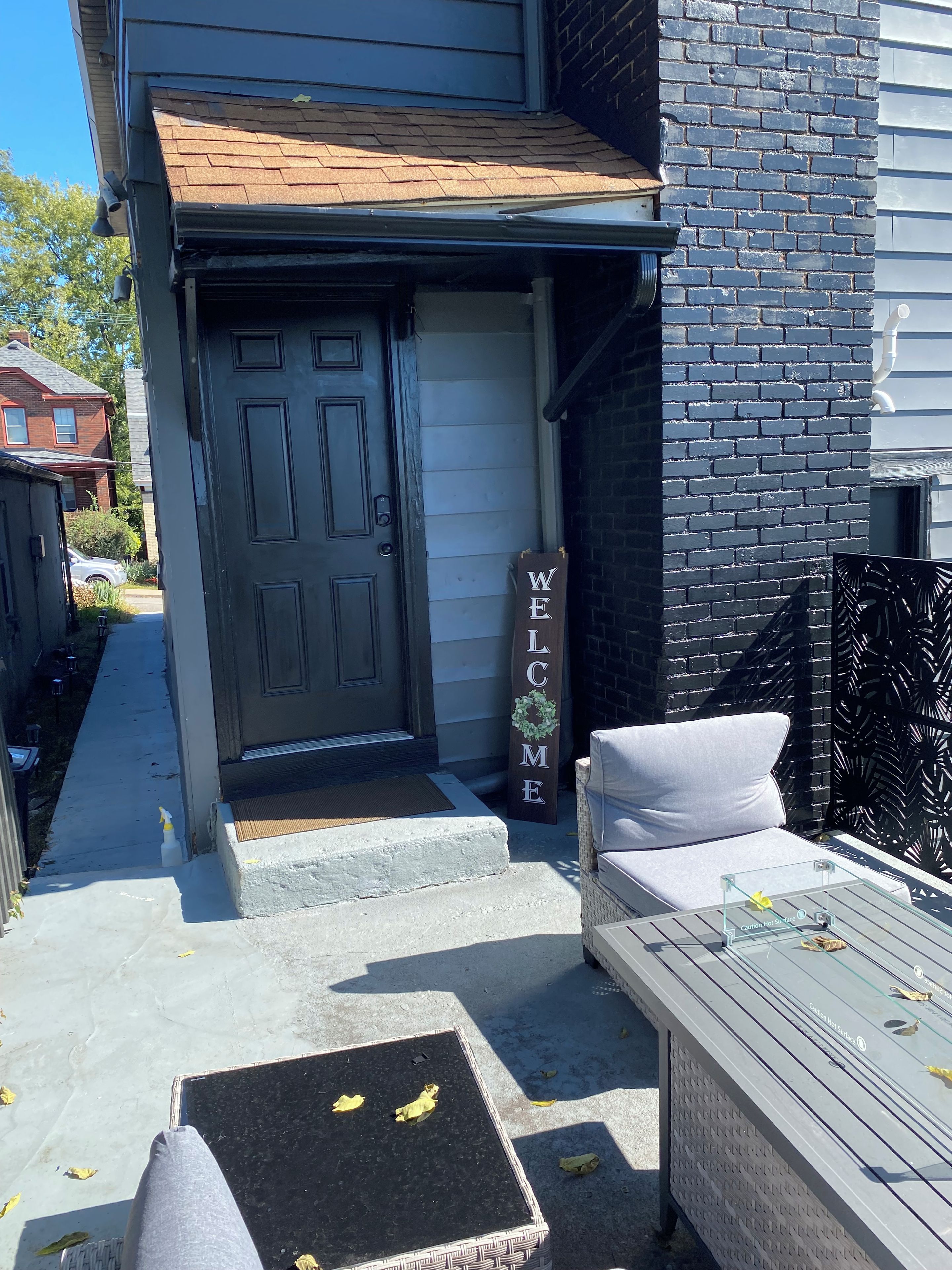 A residential entryway with a dark door, a welcome sign, outdoor seating, and a concrete step leading to the entrance.