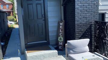 A residential entryway with a dark door, a welcome sign, outdoor seating, and a concrete step leading to the entrance.