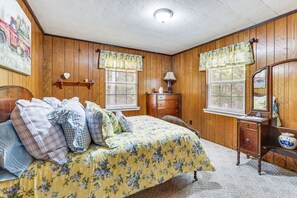 A bedroom with wooden paneling, a bed with floral bedding, two windows with curtains, a dresser, and a mirror. - Cozy Soddy-Daisy Home w Fire Pit & Porch (Soddy-Daisy)