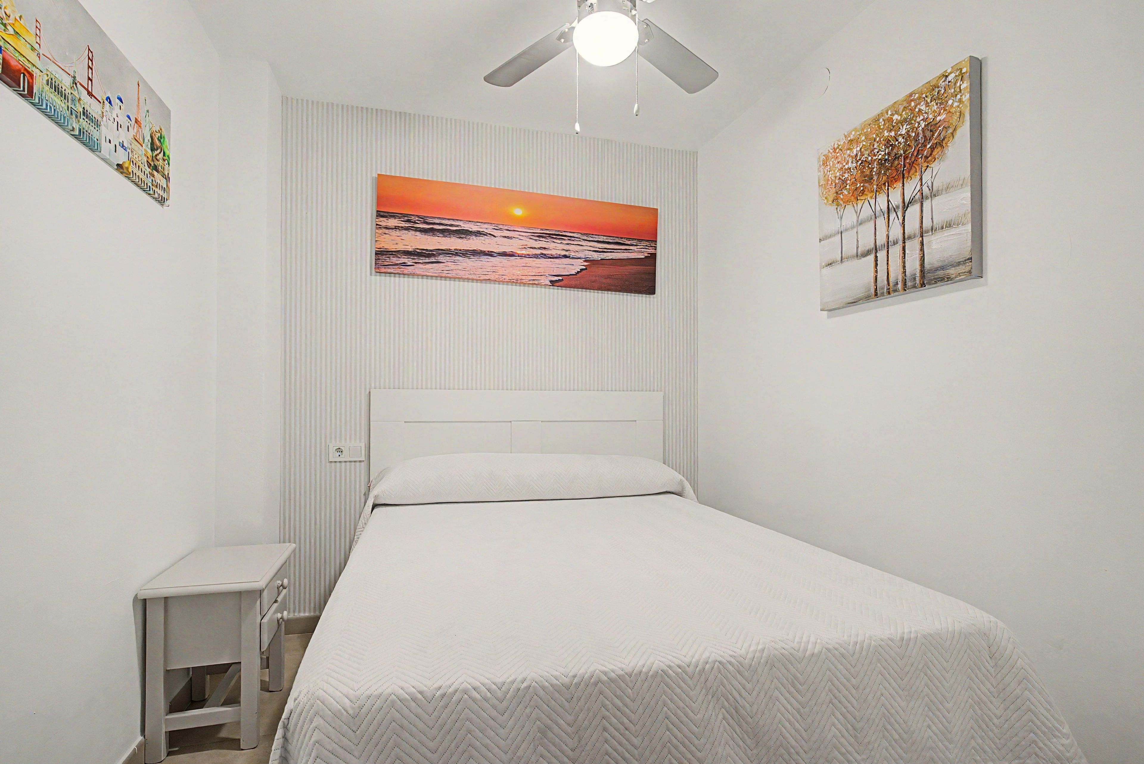 Ein Schlafzimmer mit einem Bett, einem Deckenventilator, einem Sonnenuntergangsbild und einem kleinen Nachttisch.