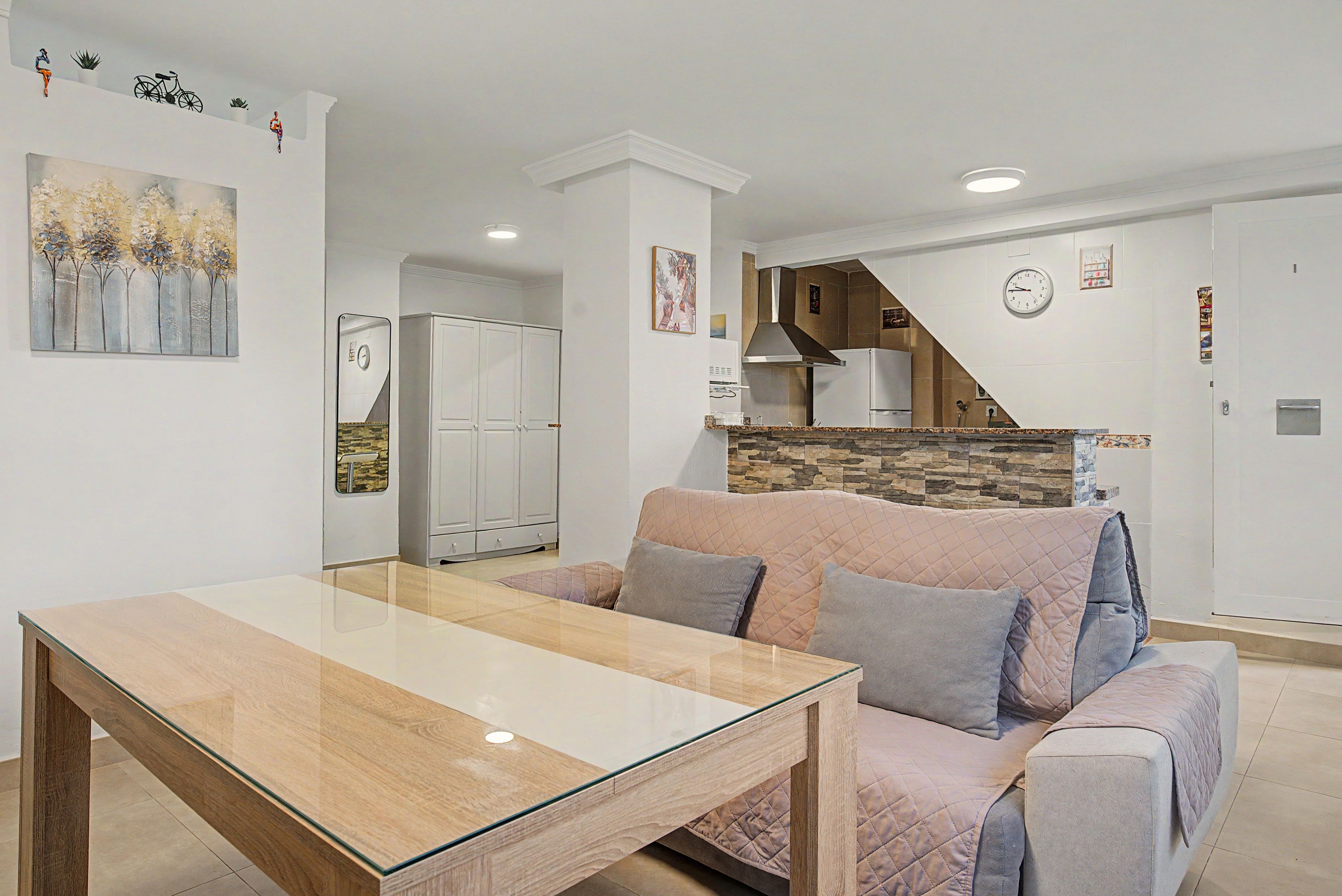 Een moderne woonkamer met een eettafel met glazen blad, een bank met kussens en een keuken met een fornuis en een koelkast.