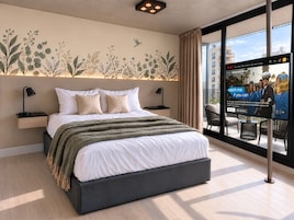 A modern hotel room with a large bed, a wall-mounted TV, and a balcony with a view of the city.
