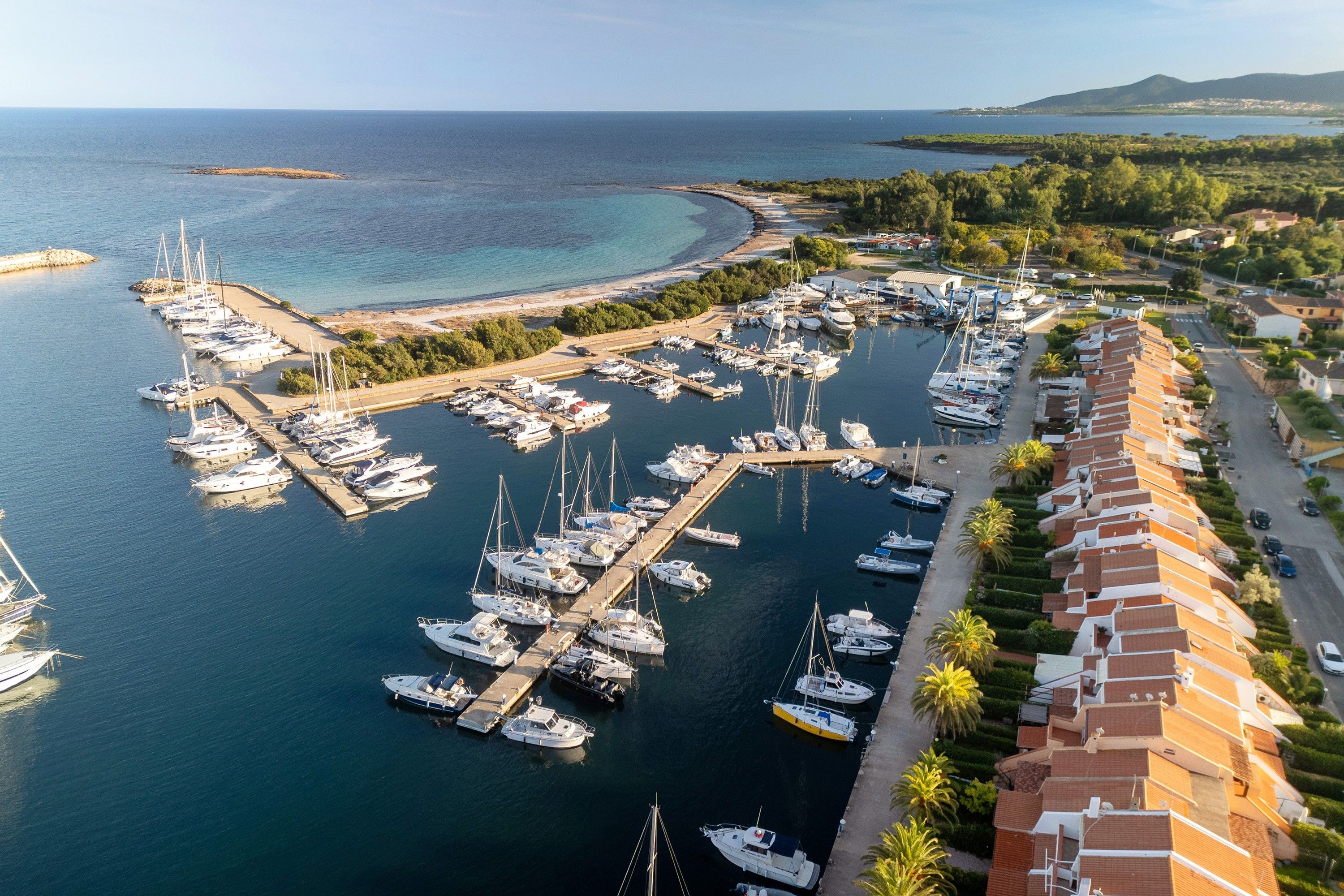Un puerto deportivo con numerosas embarcaciones amarradas, rodeado por un paisaje costero con edificios y vegetación.