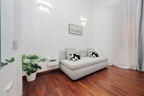 Ein modernes Wohnzimmer mit einem grauen Sofa, einem kleinen weißen Beistelltisch, einer Topfpflanze und einem gerahmten Bild an der Wand.
