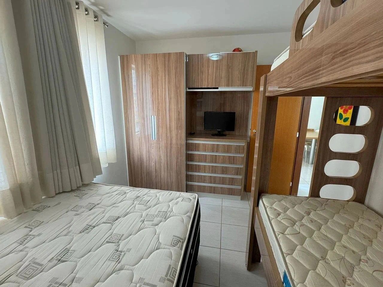 Quarto com cama de solteiro e beliche, guarda-roupa, televisão e escrivaninha.