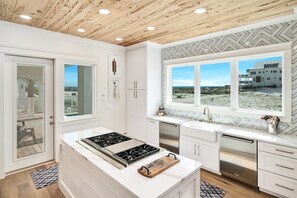 Una cocina moderna con armarios blancos, una isla central con estufa y vistas a la playa a través de amplios ventanales.