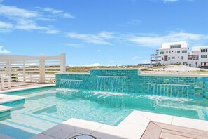 A swimming pool with a waterfall feature, surrounded by a white fence and a modern building in the background. - Amberjack | 7BR | Heated Pool & Spa | Sleeps 16 (Inlet Beach)