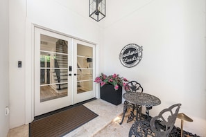 A white exterior door with a glass panel, a black planter with pink flowers, and a metal table with chairs. - Beachwalk Landing 6-Seater Golf Cart | Beach 3–5 Min | Sleeps 8 (Miramar Beach)