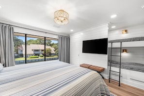Quarto moderno com cama grande, beliche, televisão e vista para uma casa através de uma porta deslizante.