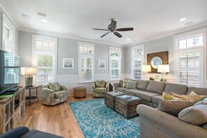 A spacious living room with a large sofa, a coffee table, and a television. There are multiple windows with blinds, a ceiling fan, and a decorative mirror. - Blue Grouper | Pool | Bikes | Games | 30A (Seagrove Beach)