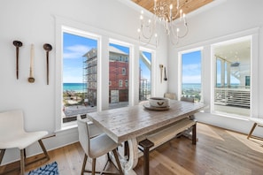 A room with a large wooden table, a white chair, and a view of the beach and construction through large windows. - Coastal View | Gulf Views | Pool & Hot Tub (Inlet Beach)