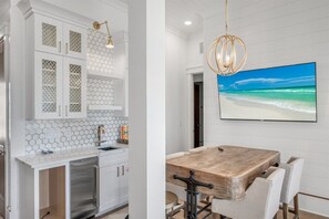 Eine moderne Küche mit weißen Schränken und einer Rückwand aus sechseckigen Fliesen, ein Essbereich mit Holztisch und Stühlen sowie ein großer Flachbildfernseher, der eine Strandansicht zeigt.