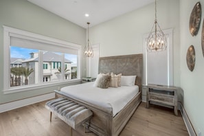 A bedroom with a large bed, a bench, a nightstand, and a view of houses through a window. - Coral Cay | Pool, Hot Tub & Elevator | Beach Getaway (Inlet Beach)