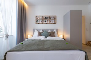 Una cama bien tendida con un edredón gris, cabecera con varios cojines, lámparas de noche y una composición de fotos en la pared.