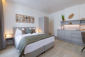Un dormitorio moderno con cama, lámparas de noche, mesitas de noche, un cuadro colgado en la pared, una planta pequeña y una zona de cocina con fregadero y armarios.