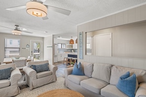 A spacious living room with a large beige sofa, two armchairs, and a coffee table. There is a ceiling fan, a kitchen in the background, and a sign that reads 'BEACH HOUSE'. - 30A Dogwood Bungalow | Walk to Beach | Pet Friendly (Santa Rosa Beach)