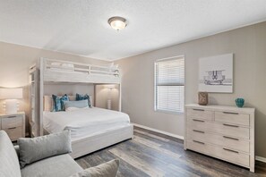 Ein Schlafzimmer mit Etagenbett, Nachttisch mit Lampe, Kommode und Fenster mit Jalousien.