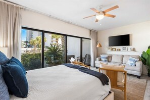 Quarto de hotel com cama, sofá, televisão e varanda com vista para palmeiras.