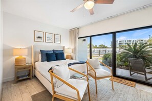 Una camera d'hotel con un letto, due sedie, un comodino, una lampada e un'ampia finestra che si affaccia all'esterno.