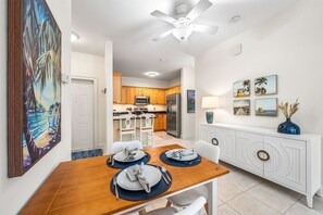 Uma sala de jantar com mesa de madeira posta para quatro lugares, cadeiras brancas, um ventilador de teto e uma cozinha ao fundo.