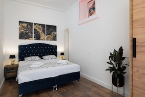 A bedroom with a dark blue bed, white bedding, and two wall-mounted lamps. There are three framed artworks above the bed and a potted plant to the right. - Luxury apartment with patio in the heart of Split (Split)