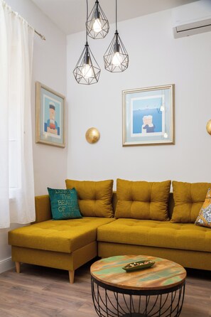 Uma sala de estar com um sofá cor mostarda, uma mesa de centro redonda e duas luminárias pendentes.