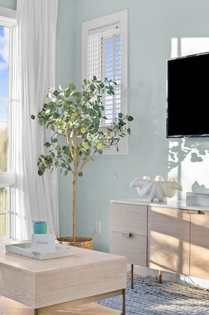 Ein Zimmer mit einem Holztisch, einer Topfpflanze, einem an der Wand montierten Fernseher und einem Fenster mit weißen Vorhängen.