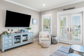 A living room with a flat-screen TV, a white armchair, a round ottoman, a patterned rug, and a cabinet with decorative items. - Sandy Knickers | Pool | Gulf Views | Beach Access (Seacrest)