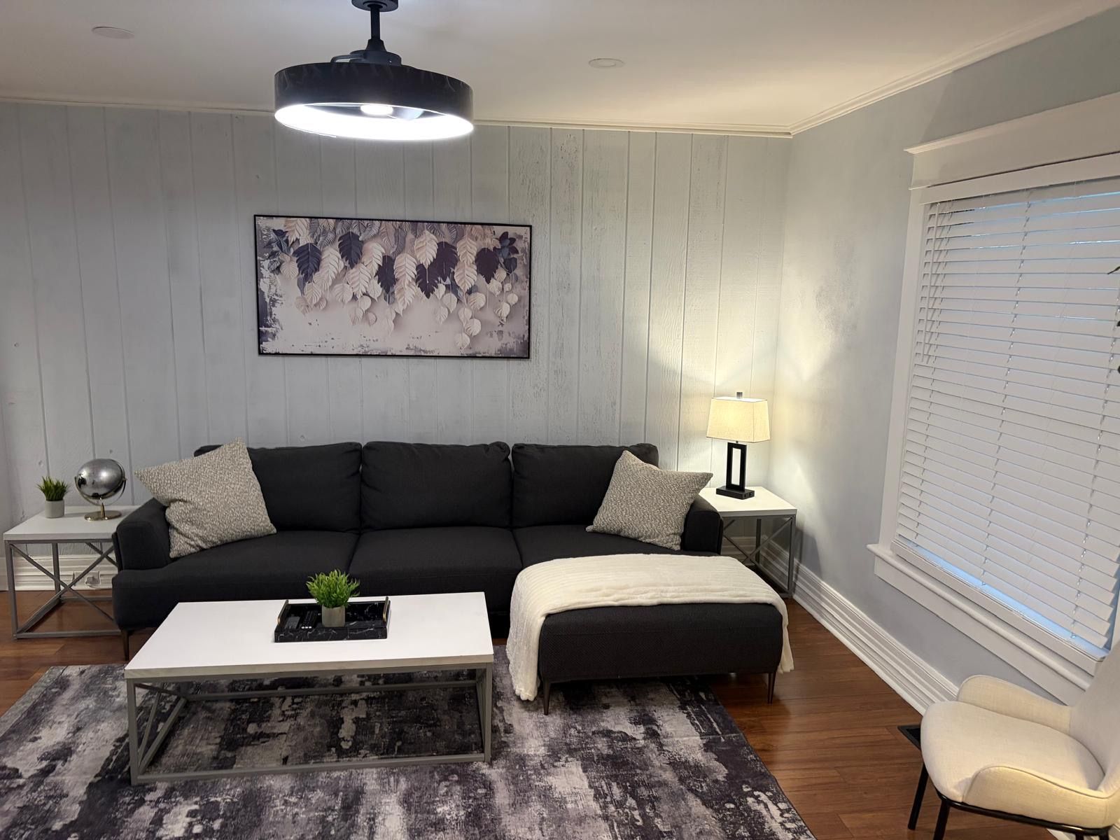 A living room with a black sofa, a white coffee table, a grey rug, a floor lamp, and a window with blinds.