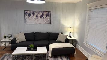 A living room with a black sofa, a white coffee table, a grey rug, a floor lamp, and a window with blinds.