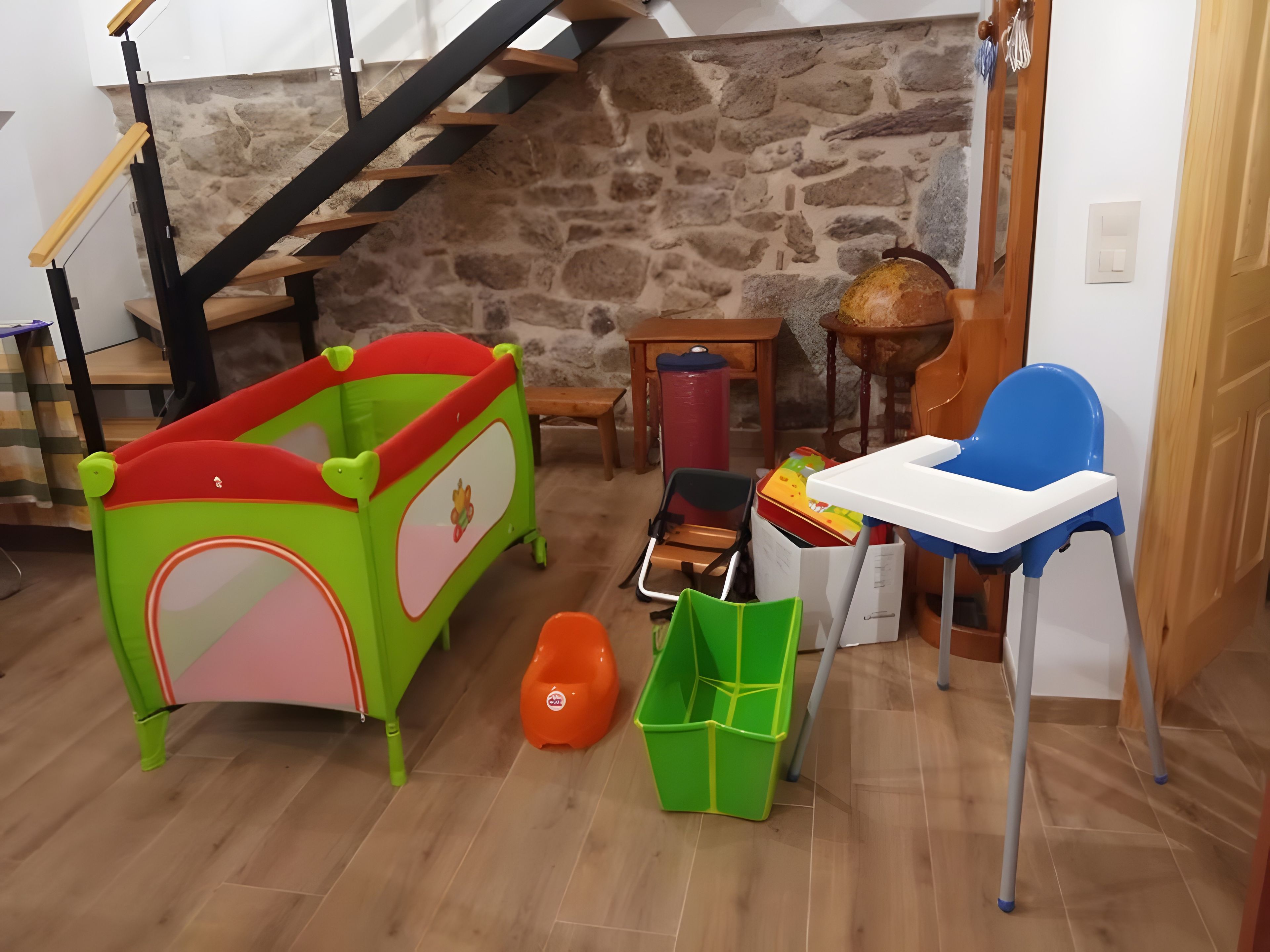 Habitació amb una escala de fusta, un parc infantil verd, una cadireta alta blava i una taula petita.