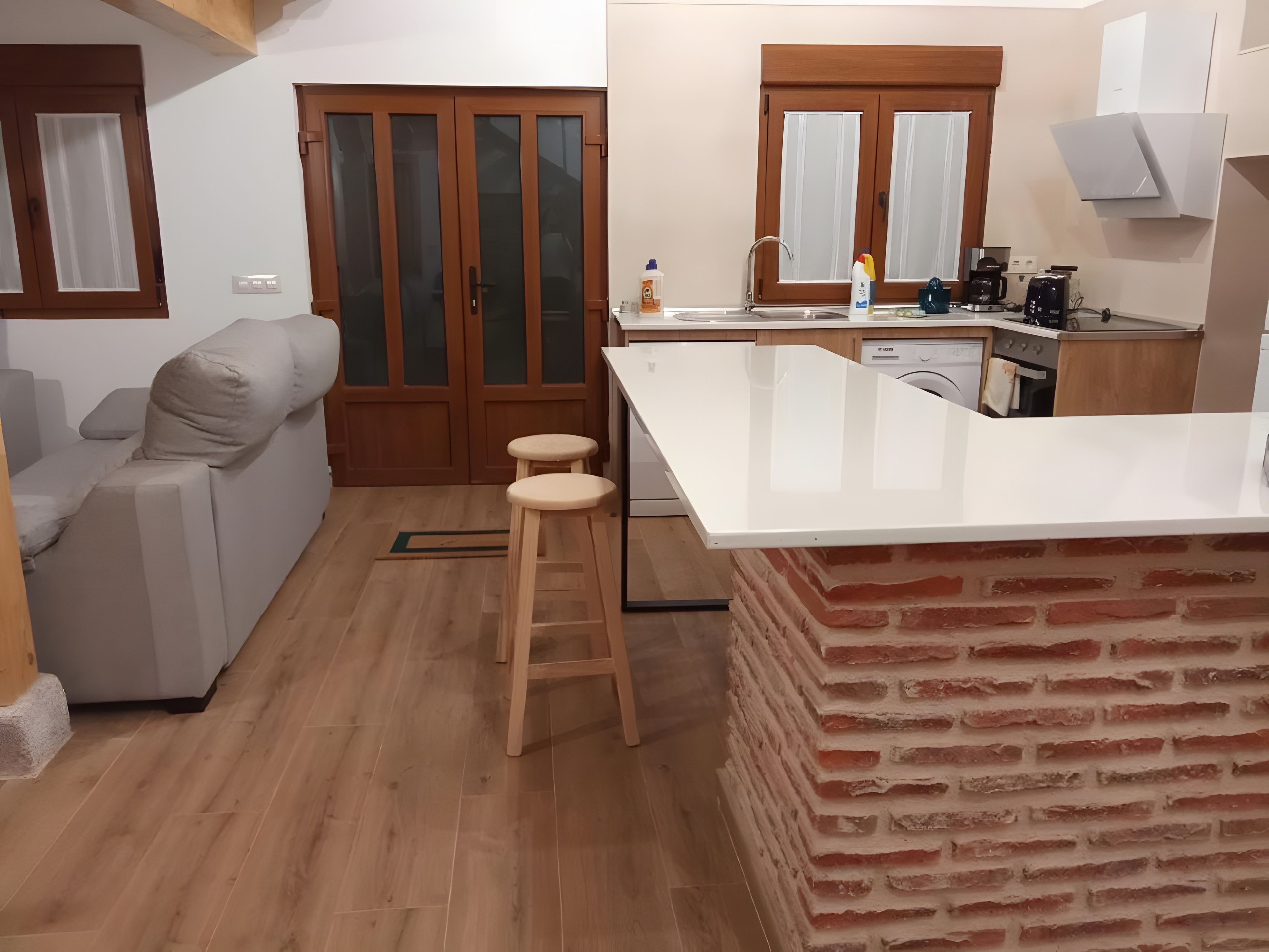 A compact kitchen with a white countertop, wooden stools, a brick accent wall, and a sliding glass door leading to a living area.