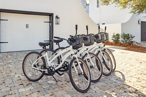 A row of white bicycles with baskets parked on a cobblestone driveway in front of a white building with a garage door. - Stepping Stone Main & Carriage | Pool | Sleeps 14 (Inlet Beach)