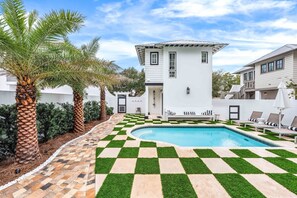 A pool area with a checkered pattern, palm trees, and a white building with a balcony. - Stepping Stone Main & Carriage | Pool | Sleeps 14 (Inlet Beach)