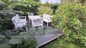 A wooden deck with white outdoor furniture, surrounded by lush greenery and fruit-bearing bushes.