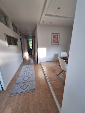 A hallway with a wooden floor, a patterned rug, a white chair, and a painting on the wall.