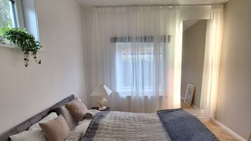 A bedroom with a bed, a bedside table, a lamp, a window with curtains, and a plant on the window sill.