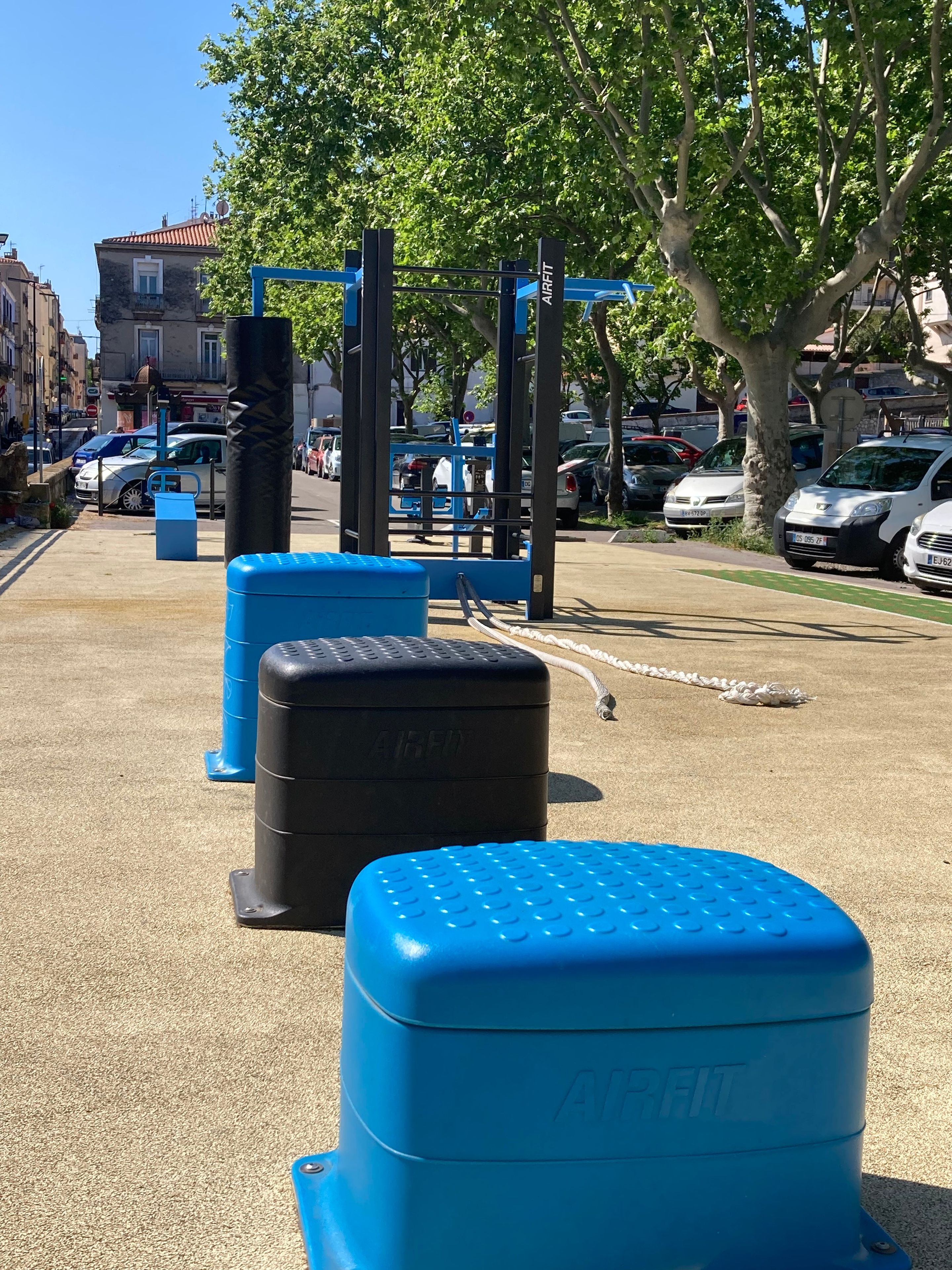 Des appareils de musculation en plein air, notamment une barre de traction et des marches bleues pour les exercices, dans un environnement urbain, avec des voitures garées et des bâtiments en arrière-plan.