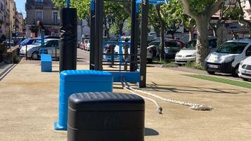 Des appareils de musculation en plein air, notamment une barre de traction et des marches bleues pour les exercices, dans un environnement urbain, avec des voitures garées et des bâtiments en arrière-plan.