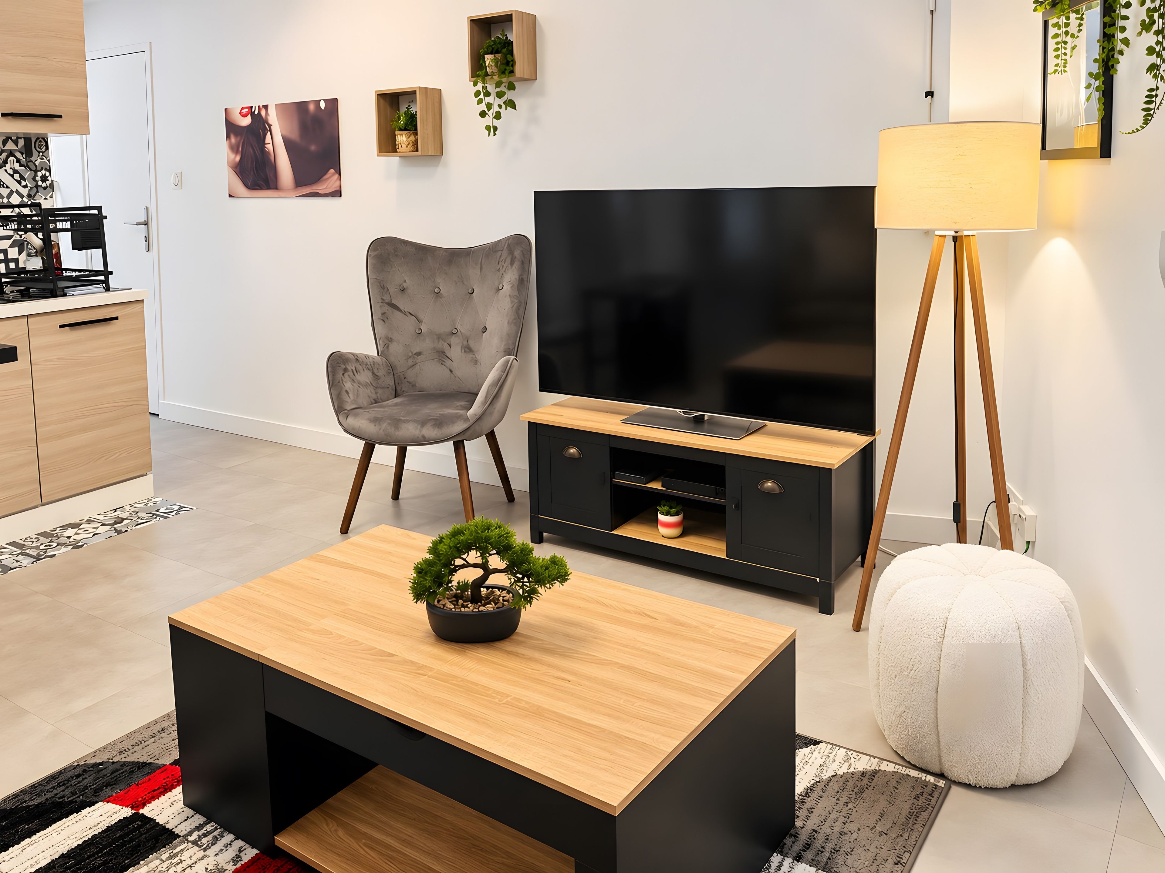 Een moderne woonkamer met een flatscreen televisie, een grijze fauteuil, een houten salontafel met een bonsaiboom, een zwarte tv-meubel, een vloerlamp en een witte poef.