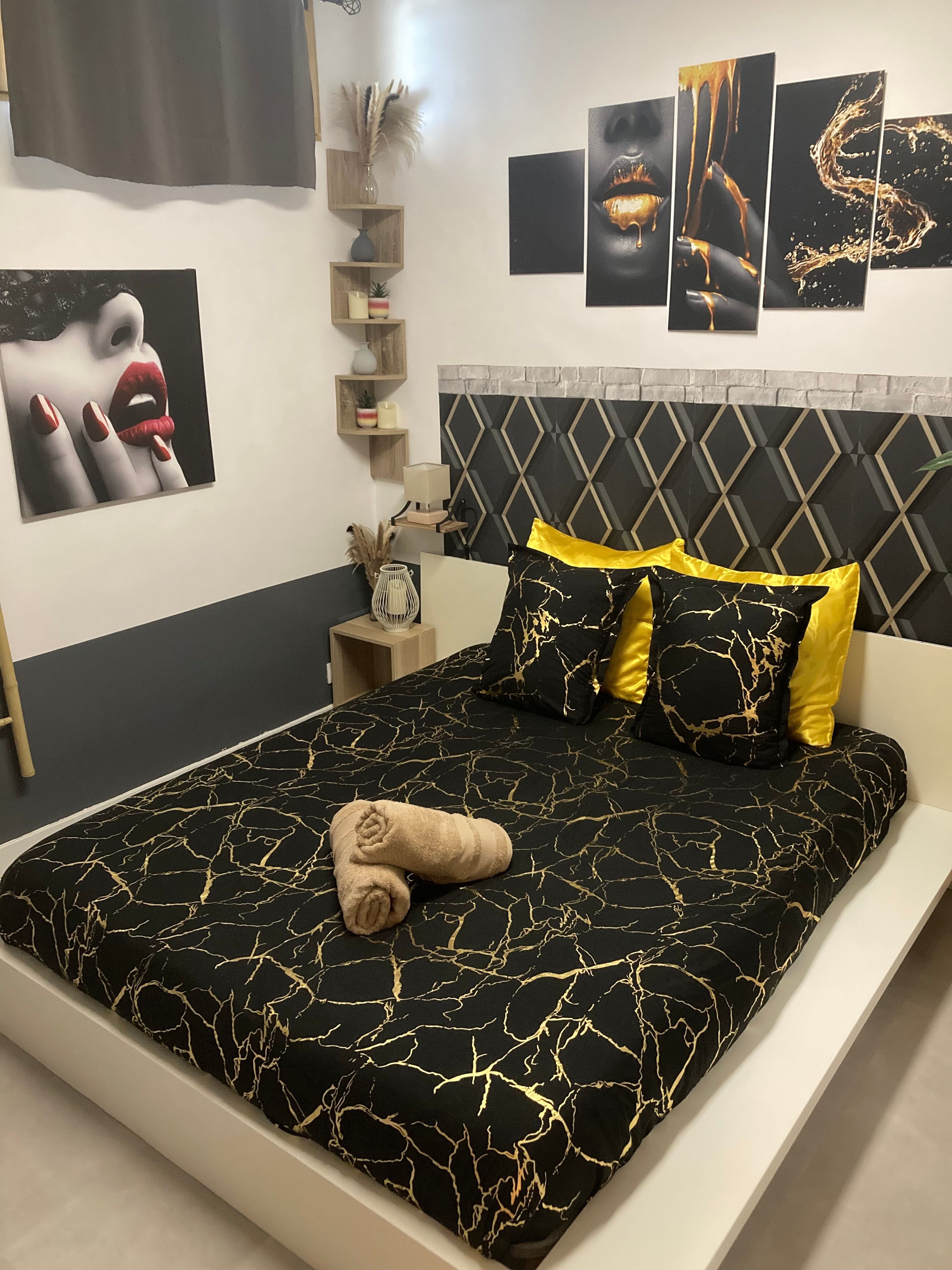 Een moderne slaapkamer met een zwart-gouden bedsprei, een hoofdbord met een geometrisch ontwerp en een nachtkastje met een lamp en decoratieve objecten.