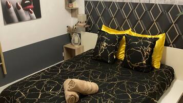 Een moderne slaapkamer met een zwart-gouden bedsprei, een hoofdbord met een geometrisch ontwerp en een nachtkastje met een lamp en decoratieve objecten.