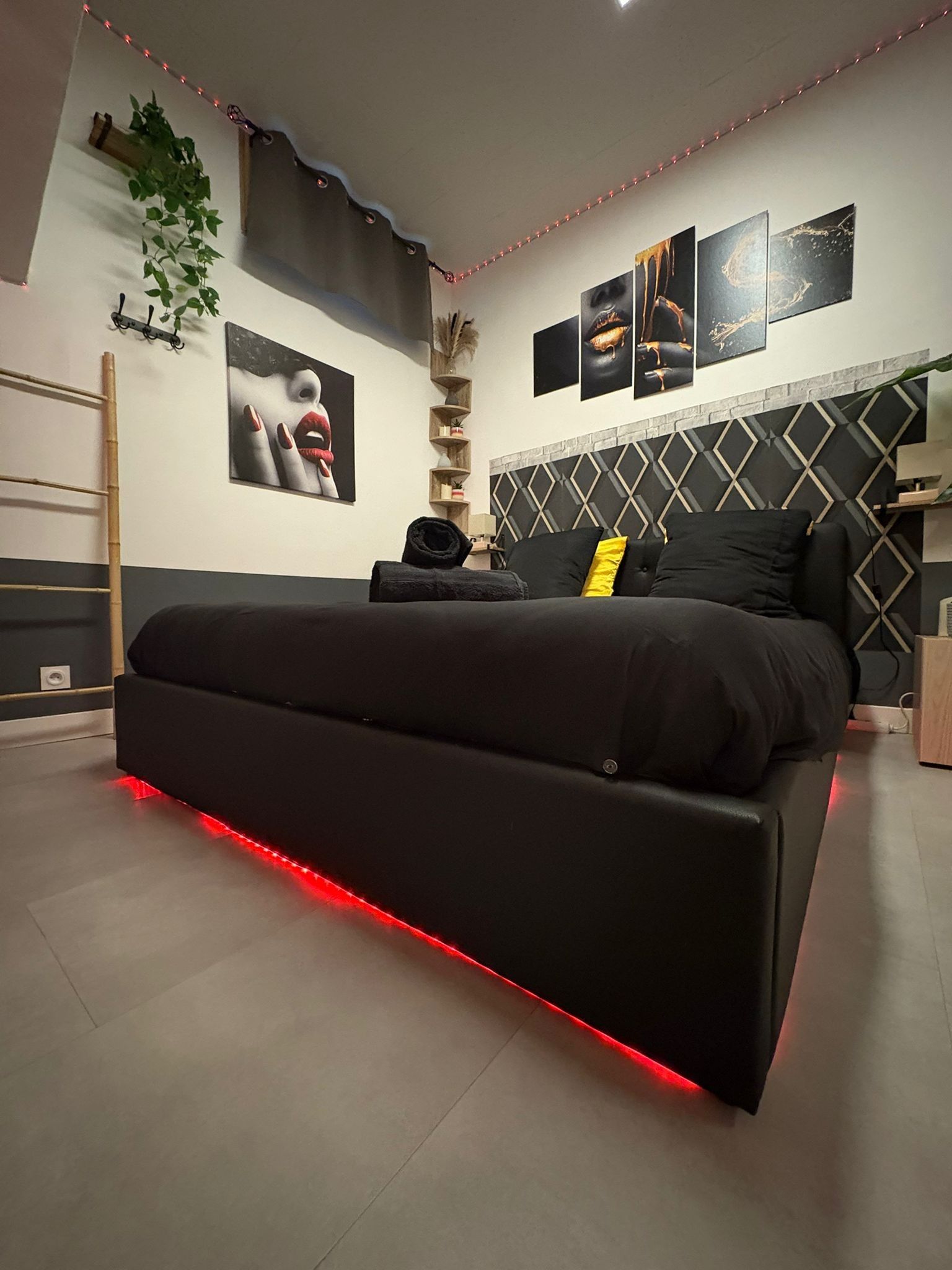 Une chambre moderne avec un lit noir, un éclairage rouge sous le lit, une tête de lit à motifs géométriques et des œuvres d’art accrochées au mur.