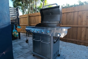 Un barbecue extérieur en acier inoxydable, doté de quatre brûleurs et d’un brûleur latéral, installé sur une terrasse en béton avec une clôture en bois en arrière-plan.