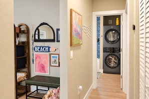 A laundry room with two stacked front-loading washers and dryers, a wooden bench, and a wall-mounted shelf with various items. - This Must Be The Place (Redington Shores)