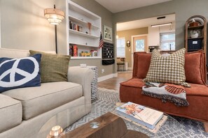 A living room with a beige sofa, a red armchair, and a coffee table with books. There is a bookshelf with various items and an American flag. - This Must Be The Place (Redington Shores)