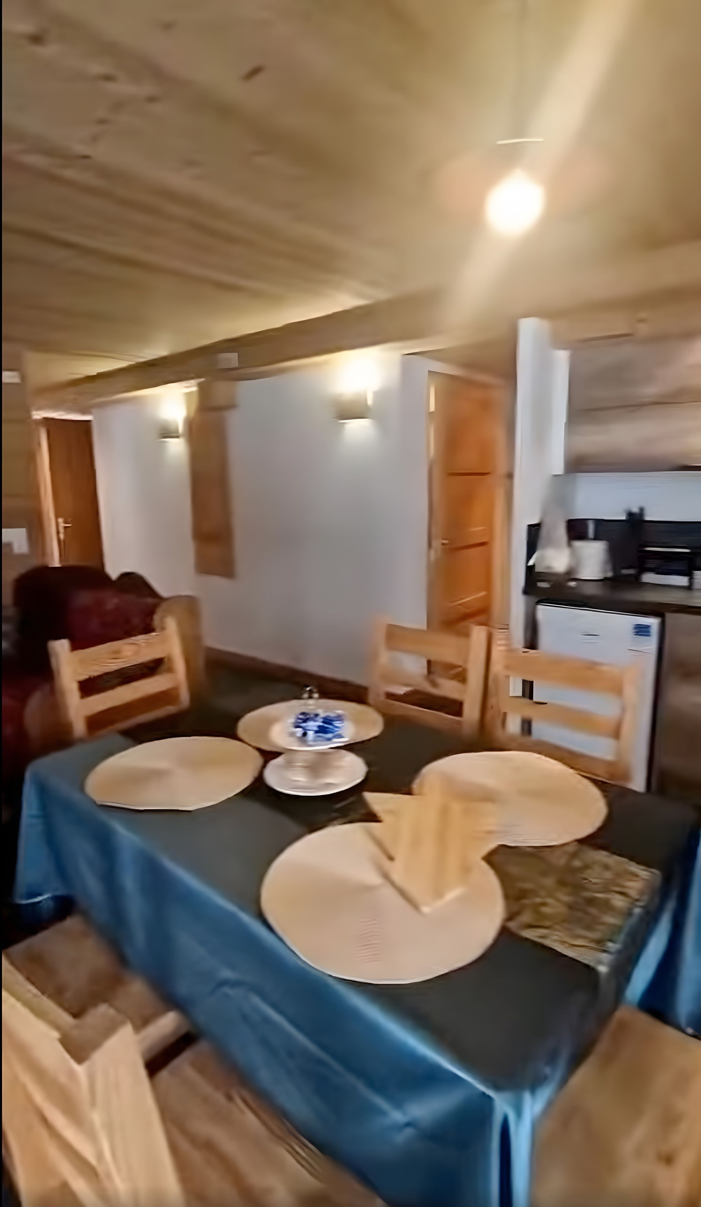 Un comedor con una mesa lista para cuatro personas, sillas de madera y una zona de cocina con refrigerador y lavaplatos.