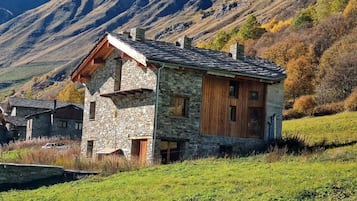 A stone house with a wooden exterior, surrounded by a grassy hillside and mountains in the background.